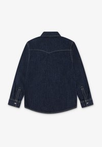 Mørk denimskjorte med lange ermer, knappevridere og krage. Har yoke-design og synlige sømdetaljer.