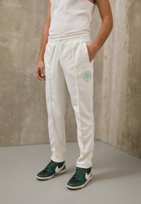 Vitaatletiska byxor med elastisk midja och sidfickor. Har en grön cirkulär logotyp. Kombineras med svarta och gröna sneakers.