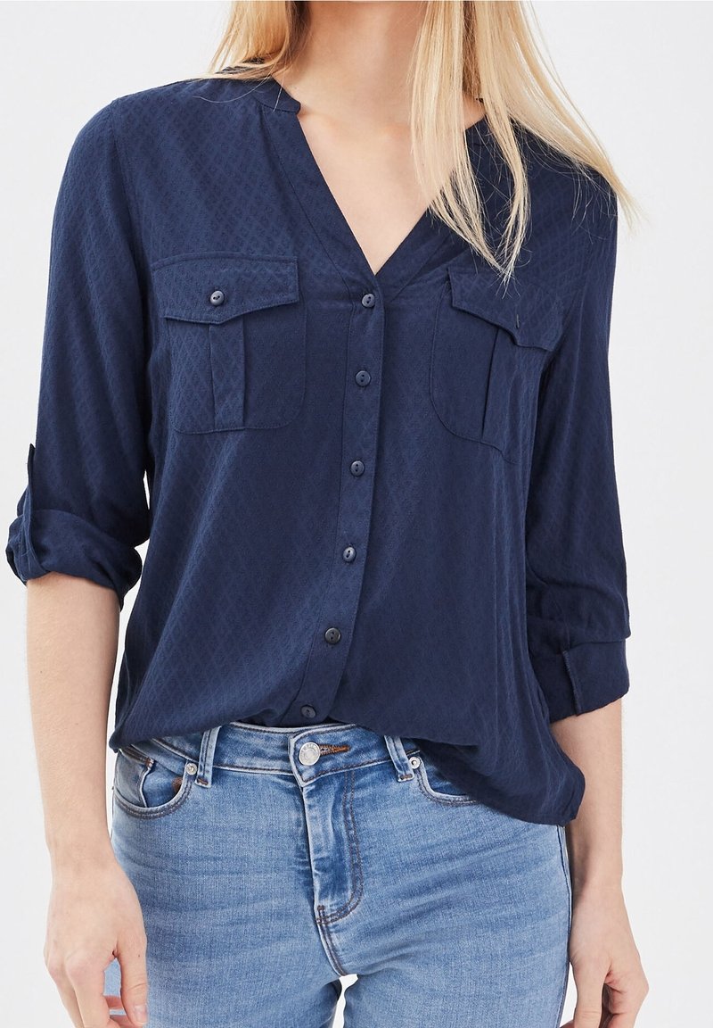 Femme portant une chemise bleu marine boutonnée aux manches retroussées et un jean bleu clair, debout devant un fond uni.