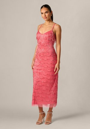 Kobieta w różowej midi sukience z koralikami na cienkich ramiączkach i srebrnych sandałach na wysokim obcasie, stojąca na jednolitym tle.