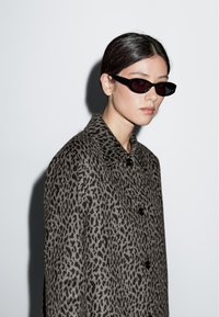 Cappotto leopardato in grigio e nero con colletto e bottoni. Occhiali da sole neri e angolari con lenti scure. Sfondo bianco semplice.
