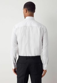 Weißes gemustertes Langarmhemd mit Button-Down-Kragen, kombiniert mit schwarzen Hosen. Merkmale sind ein dezentes strukturiertes Design und knöpfbare Manschetten.
