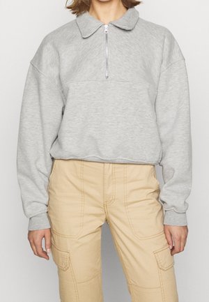 Personne portant un sweat-shirt à fermeture éclair quart de zip gris clair et un pantalon cargo beige, debout devant un fond clair uni.