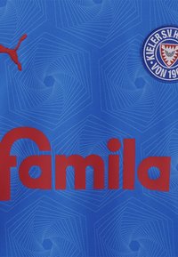 Mėlyna sportinė marškinė su geometriniu raštu, raudonas "Puma" logotipas ir ryškus raudonas "famila" tekstas, su „Kieler S.V." emblema ant krūtinės.