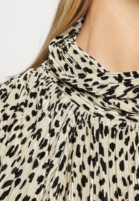 Blusa alta nera e crema con design a plissettature, caratterizzata da un motivo di macchie nere su un tessuto testurizzato e luccicante.
