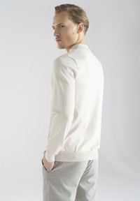 Jasno beżowy sweter dzianinowy z długimi rękawami, ściągaczami przy mankietach i u dołu. Nosi się go z szarymi spodniami w kratę, prezentując tylny widok stroju.