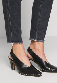 Tacones de charol negros con tiras traseras y punta afilada, tachuelas plateadas a lo largo del borde y un tacón resistente; combinados con jeans de mezclilla oscuros deshilachados.