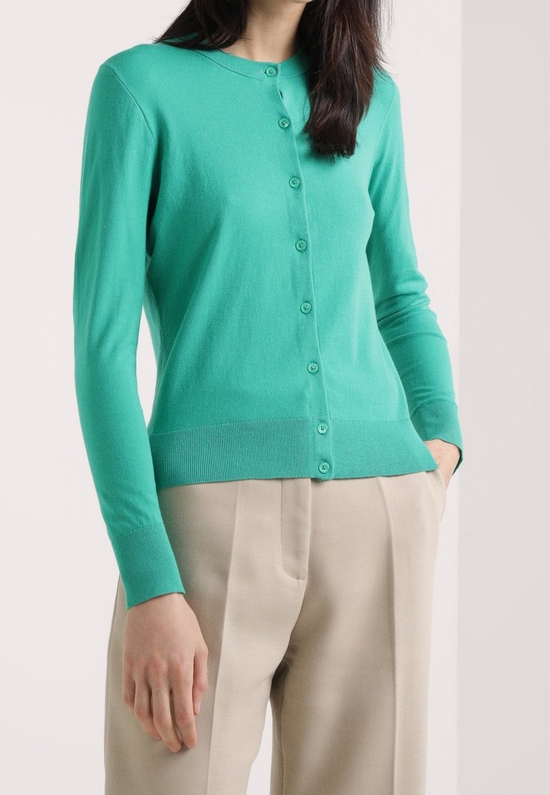 Femme portant un cardigan turquoise moulant boutonné et un pantalon beige taille haute, debout avec une main dans la poche.