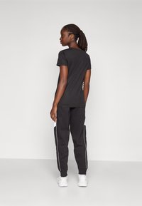 Camiseta preta de mangas curtas, combinada com calças de jogging pretas com painéis laterais brancos e punhos elásticos. O modelo usa sapatilhas de desporto brancas.