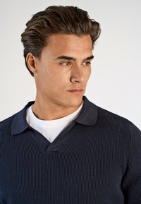 Marineblaues, geripptes Polokleid mit kontrastierendem Kragen und V-Ausschnitt, über einem weißen Hemd getragen. Glatte Textur und figurbetonte Passform.