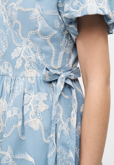 Modré šaty s bílou výšivkou mořských živočichů a mušlí, uvázané mašlí v pase, s odhalenou paží a nafouklým rukávem.