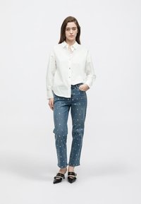 White button-up shirt, blue denim jeans with floral embroidery, black heeled shoes, standing on a neutral background.