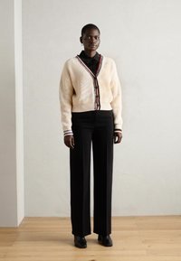 Cardigan a coste crema con bordi neri e rossi, abbinato a pantaloni larghi neri e scarpe nere, su uno sfondo chiaro.