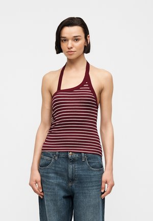 Νεαρή γυναίκα που φοράει αμάνικο μπορντό και λευκό ριγέ τοπ με halter και μπλε τζιν, στέκεται μπροστά σε απλό φωτεινό φόντο.