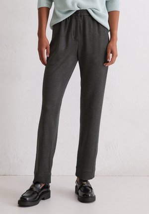 Persoon draagt donkergrijze rechte broek, zwarte loafers en een lichtmintgroene trui, staand tegen een effen witte muur.