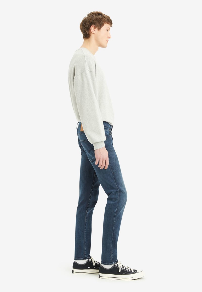 Grauer Sweatshirt mit gerippten Bündchen, slim-fit dunkelblaue Jeans und schwarze Sneaker mit weißen Sohlen. Modell in Seitenansicht gezeigt.