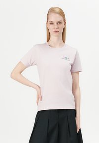 Šviesiai rožinė medvilninė marškinėliai su trumpomis rankovėmis, apvaliu kaklu ir siuvinėjimu, kuriame pavaizduoti spalvingi gėlėti raštai. Modelis pozuoja, laikydamas rankas ant klubų.