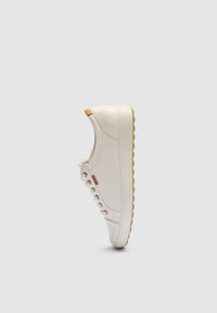 Zapatilla blanca de caña baja con suela beige, ojetes metálicos y detalle de cuero marrón claro en el talón, mostrada de perfil sobre fondo gris claro.
