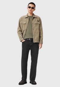 Beige corduroy jack met twee borstzakken, olijfgroen shirt eronder, zwarte jeans en zwarte schoenen. Model draagt een zonnebril.
