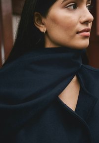 Woman with dark hair wearing small gold hoop earrings and a dark blue coat with a high collar, looking to the right.