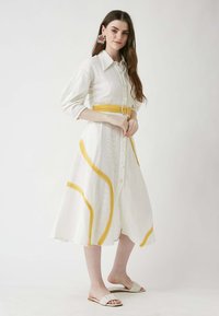 Weißes Hemdkleid mit gelben Akzenten, semitransparentem strukturiertem Stoff, Dreiviertelärmeln und einem passenden Gürtel. Flache beigefarbene Sandalen.