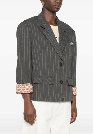 Persona che indossa un blazer grigio scuro a righe sottili con due bottoni e polsini rosa decorati, abbinato a pantaloni bianchi.