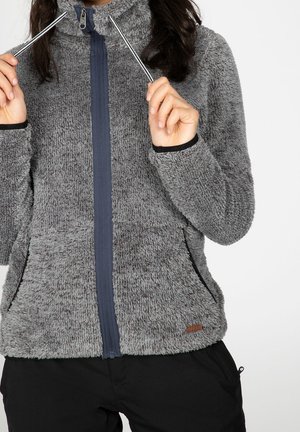 Femme ajustant les cordons d'un blouson zippé gris en tissu pelucheux porté sur un pantalon noir, sur fond blanc.