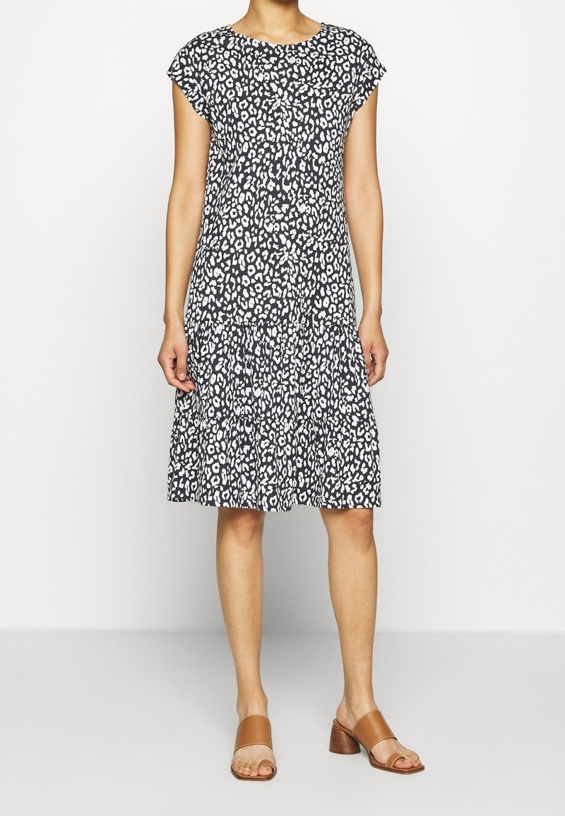 Kvinna iklädd en kortärmad klänning med svart och vitt leopardmönster, i kombination med beige öppna klacksandaler, står framför en vit bakgrund.