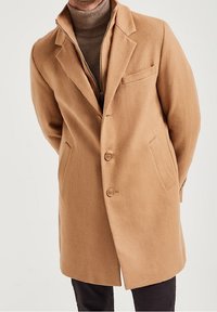 Kamelullblandad kappa med notchkrage, knappar fram, sidfickor och bröstficka, lagd över en ribbad beige turtleneck.