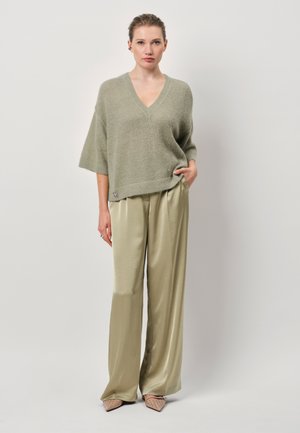 Pullover en maille vert clair, surdimensionné, avec un col en V ; associé à un pantalon en satin beige brillant, taille haute et à jambes larges. Chaussures beiges à bout pointu.