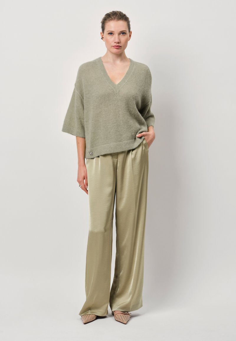 Lichtgroene, oversized gebreide trui met een V-hals; gecombineerd met hooglopende, wijde, glanzende beige satijnen broeken. Puntaarde beige schoenen.