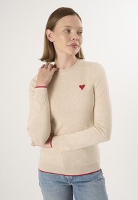 Beige Strickpullover mit Rundhalsausschnitt, der auf der Brust eine kleine rote Herzstickerei und kontrastierende rote Ärmelabschlüsse und einen Saum aufweist.