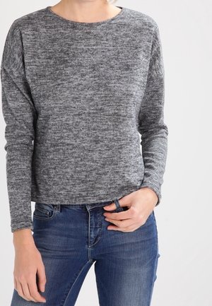 Personne portant un pull gris chiné à manches longues et un jean bleu, une main tenant la ceinture du jean.