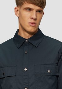Ciemnoniebieska koszula z zapięciem na guziki, posiadająca dwie kieszenie na piersi oraz srebrne metalowe napy, wykonana z lekkiego materiału o gładkiej fakturze.