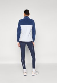 Fleecejakke i marineblå og hvit med høy krage; kombinert med mørke leggings med hvite detaljer og logo; grå treningssko.