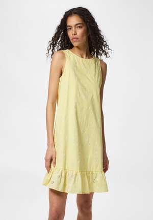 Femme aux cheveux bouclés et foncés portant une robe jaune à fleurs sans manches avec un ourlet volanté, debout devant un fond blanc uni.