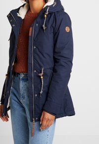 Marinblå parka med huva klädd i konstpäls, justerbara dragsnören och träknappar. Har dragkedjestängning och sidofickor.