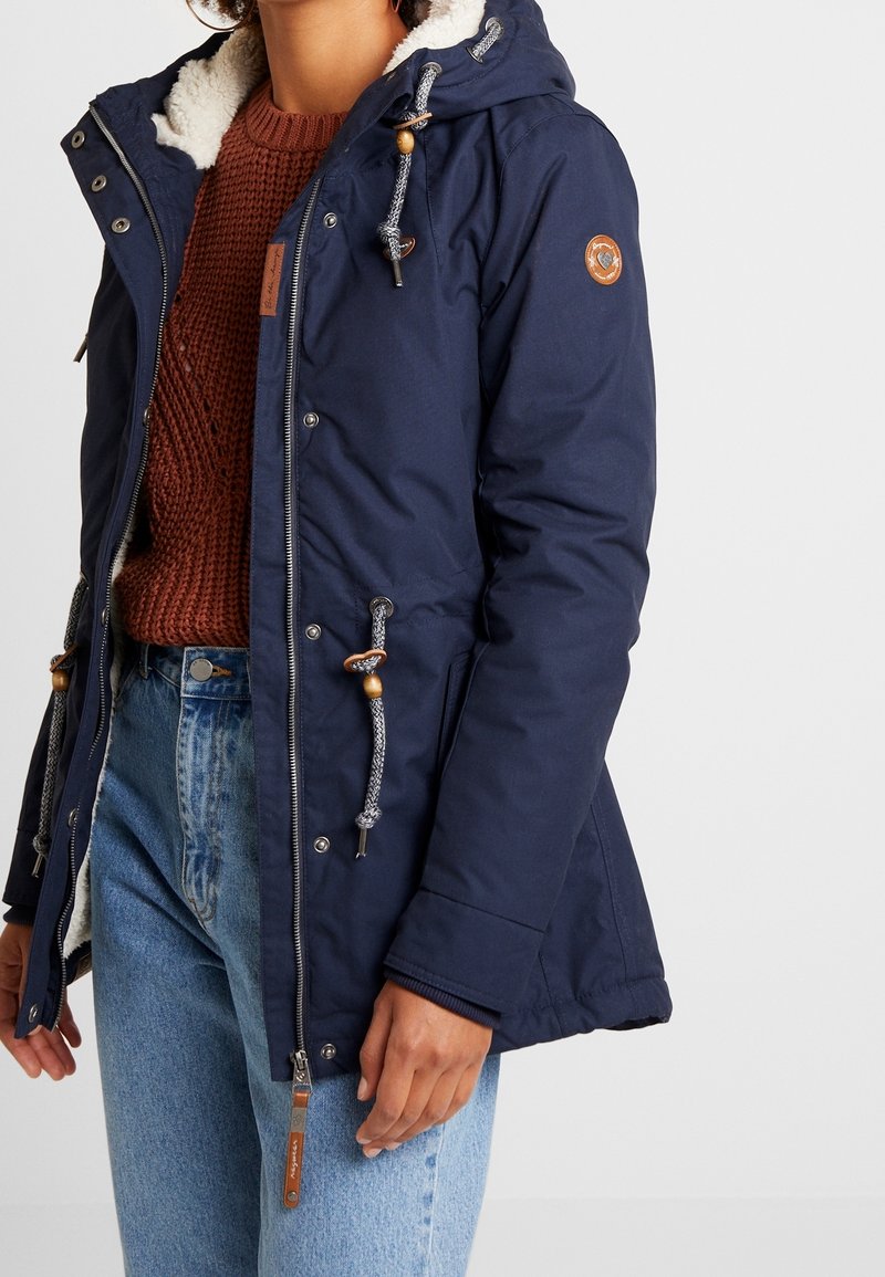 Marinblå parka med huva klädd i konstpäls, justerbara dragsnören och träknappar. Har dragkedjestängning och sidofickor.