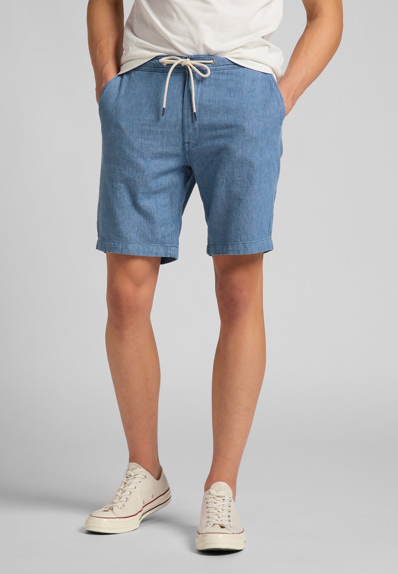 Pantalones cortos de chambray azul con cintura ajustable, bolsillos laterales y dobladillo enrollado. Combinados con zapatillas blancas. Textura suave.