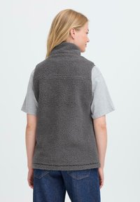 Femme portant un gilet en polaire gris sur une chemise à manches courtes gris clair et un jeans bleu, de dos contre un fond blanc uni.
