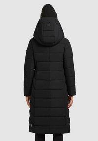 Vrouw in een lange zwarte gewatteerde winterjas met capuchon en een zwarte gebreide muts, gezien van achteren tegen een effen achtergrond.