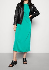 Ein türkisfarbenes Midikleid aus glattem Stoff, kombiniert mit einer schwarzen Lederbikerjacke und offenen schwarzen High-Heel-Mules.