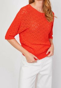 Nainen, joka käyttää kirkkaan oranssia lyhythihaista neulepaitaa, jossa on kukkakuvioinen pitsikuvio ja valkoiset housut, säätää neulepaidan helmaa.