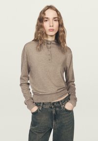 Brązowa bluzka z długim rękawem, wysokim kołnierzem i zapinaną na guziki listwą. Połączona z ciemnymi dżinsami. Miękka w dotyku, casualowy krój, minimalistyczny design.