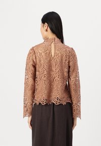 Haut en dentelle marron à manches longues et col montant, présentant des motifs floraux complexes et une ouverture en forme de trou de serrure à l'arrière.