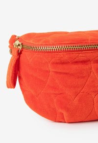 Oranje gewatteerde pouch gemaakt van zacht fluwelen stof, met een gouden rits, een ronde vorm en hartpatronen die op de buitenkant zijn gedrukt.