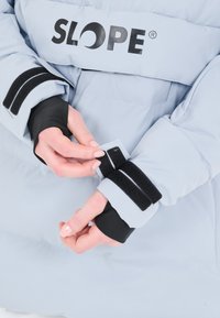 Vaaleansininen eristävä takki, jossa on mustat tarranauhakäsivarsit. Ominaisuuksina "SLOPE." Käsivarsissa on sisäpuolella mustaa kangasta paremman istuvuuden ja lämpimyyden vuoksi.