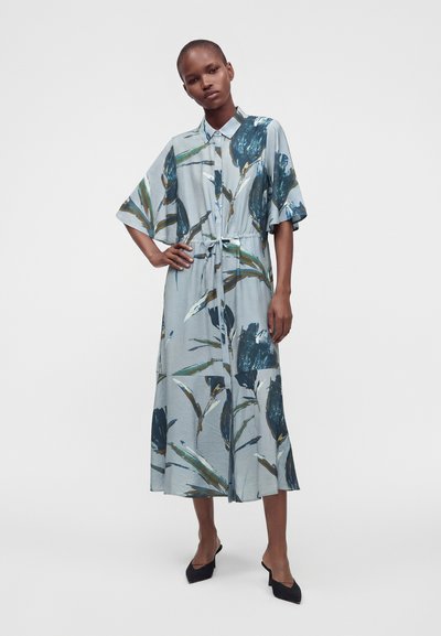 Femme portant une robe midi bleu clair avec de grands imprimés floraux foncés, des manches courtes larges, une taille cintrée et des escarpins noirs à bout pointu.