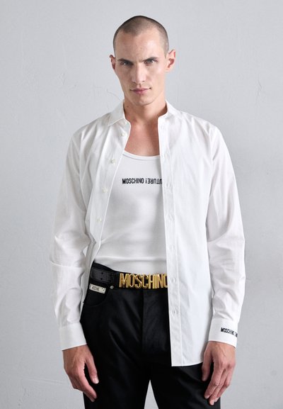 Valge särk kraega, kantud valge t-särgi peal, millel on tekst "MOSCHINO". Mustad püksid, millel on kulla värvi vöö, millel on kirjas "MOSCHINO."