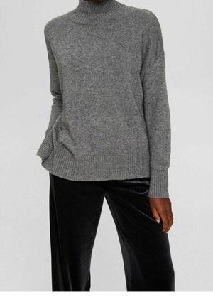Person iført en løs grå rullekravesweater og sorte fløjls brede bukser, stående med den ene hånd i lommen.